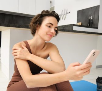 Sport and wellbeing. Portrait of fitness woman, gym instructor workout at home, sits on yoga mat and looks at her smartphone with smiling face.